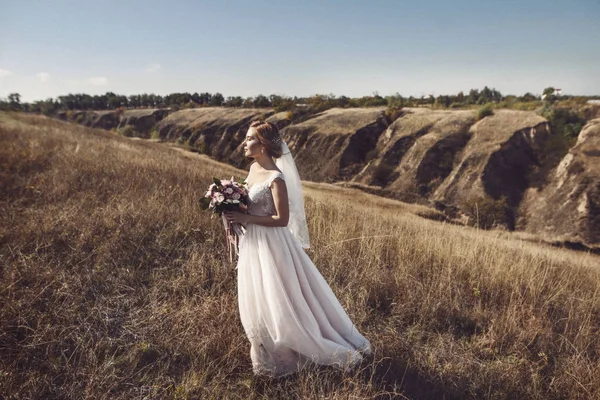 完全な長さの長い白いウェディング ドレスとベール フォレスト ブーケを保持するには、屋外自然な背景、画像の水平方向に立って 1 つ美しい官能的な若いブルネット花嫁のサイドビュー — ストック写真