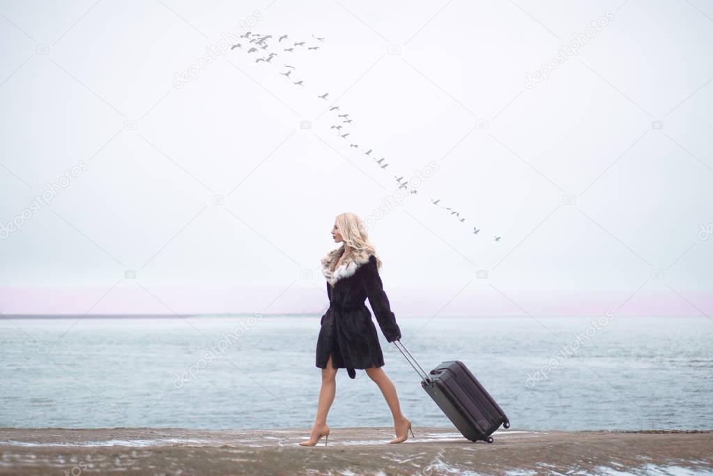 Beautiful woman in fur coat with suitcase going near sea , Birds in sky fly south, travel concept