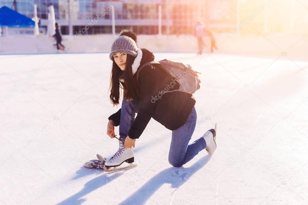 Beautiful girl having fun in winter park, ties the laces on the skates at ice rink. Enjoying nature, winter time