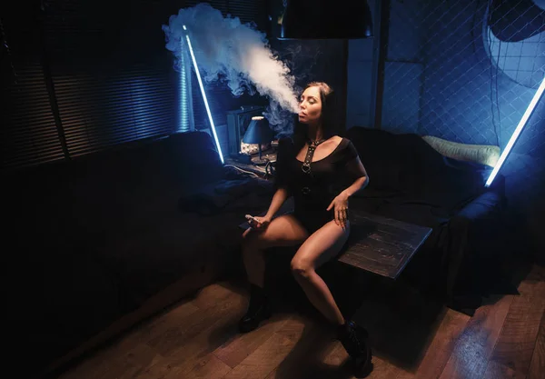 Сексуальная молодая женщина на диване, испаряющая электронную сигарету против темно-синей ба — стоковое фото