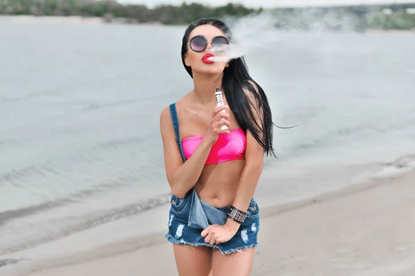 Young sexy woman with big breast vaping e-cigarette near blue water on the beach — Stock Photo, Image