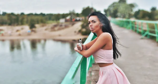 Sexy morena vapeando en puente. modelo femenino que vapea el e-líquido aromatizado de la fruta o el e-jugo con el dispositivo del vaporizador. —  Fotos de Stock