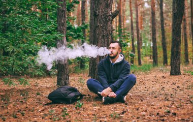 Vape adam. geri kalan ormanda sahip acımasız sakallı genç adam. Ağacın altında oturan adam. Yaşam tarzı.