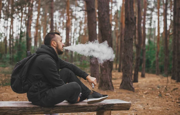 Brutal barbudo joven que descansa en el bosque y vapeo dispositivo de vapor. Estilo de vida. Concepto Vape. Descanso en el bosque — Foto de Stock