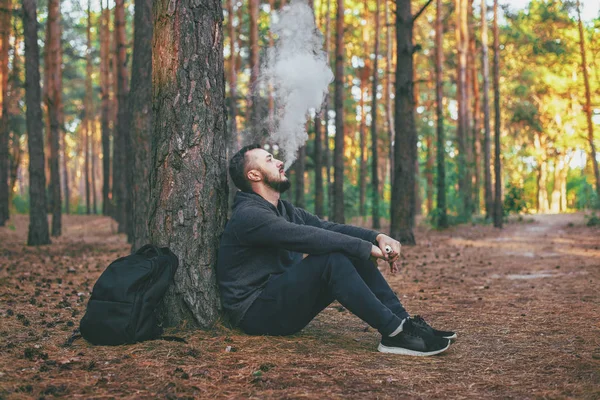 Hombre Vape. joven barbudo brutal descansando en el bosque y vapeando un cigarrillo electrónico. Estilo de vida. Concepto de Vape — Foto de Stock
