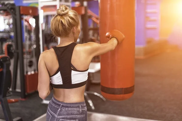 Mulher atraente perfurando um saco com luvas de boxe no ginásio. Visão traseira — Fotografia de Stock