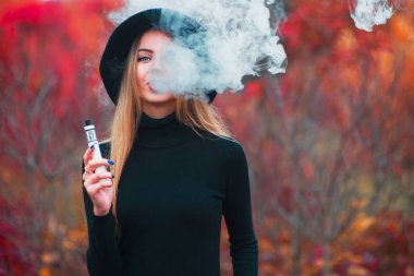 Genç güzel kız inblack şapka vaping vape aygıt vape sonbaharda açık havada bulut ile.