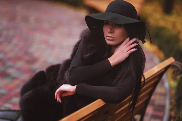 Mujer elegante en abrigo negro con un sombrero sentado en el banco en el parque de otoño —  Fotos de Stock