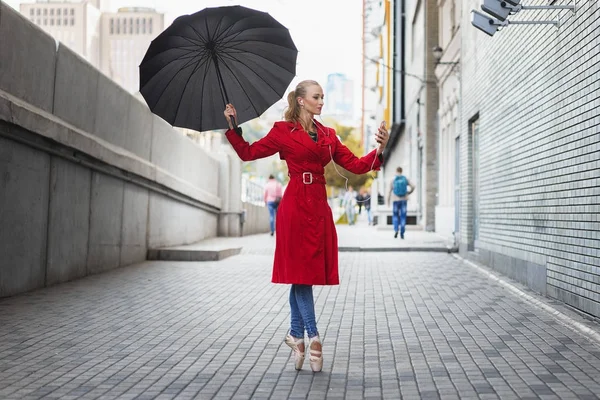 Балерина с зонтиком на городской улице. Зонтик висит в воздухе. Она стоит в балетной позе со смартфоном в руке и наушниками — стоковое фото