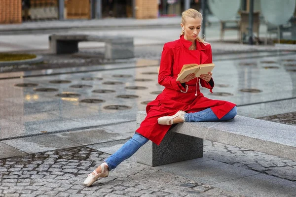 Балерина сидит на скамейке, юная красавица в красном пальто и пуантах отдыхает на улице . — стоковое фото