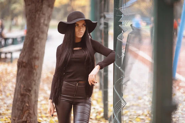 Chica en pantalones de cuero negro y sombrero cerca de la valla. Moda femenina. Estilo de vida. Cabello muy largo — Foto de Stock