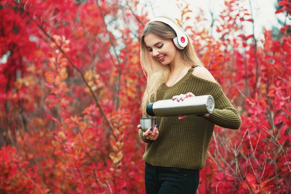 Chica sonriente sirviendo té de la taza de termo, al aire libre. Una joven bebiendo té en la taza. Viaje temático. Mujer sirviendo una bebida caliente en una taza de termo. Chica escuchar la música en haedphones —  Fotos de Stock