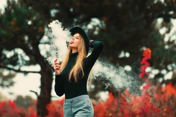 Vaping.年轻美丽的女人在黑色的帽子 vaping 户外在秋季。蒸气概念. — 图库照片