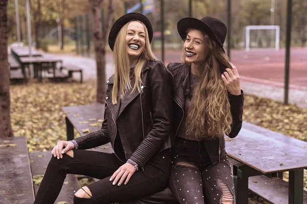 Retrato de duas belas namoradas em chapéus pretos sentado no banco rir e se divertir — Fotografia de Stock