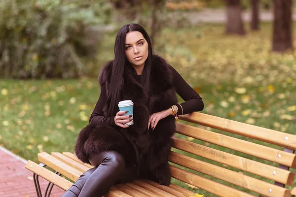 Hermosa mujer sosteniendo taza de café de papel y sentado en el banco en el parque de otoño — Foto de Stock