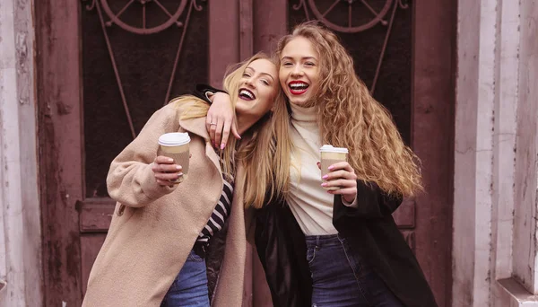 Retrato de moda al aire libre de dos jóvenes hermosas amigas tomando café. Sonriendo y yendo de compras. Besando una taza de café. Usando ropa de abrigo elegante. Maquillaje brillante —  Fotos de Stock