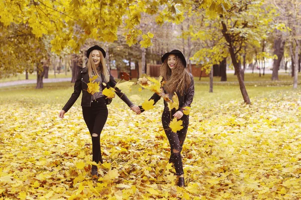 Attractive young two happy woman in hats and black clothing having fun on yeloow leaves outdoors smiling — Stock Photo, Image