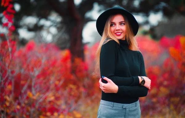 Feliz joven en el parque en el soleado día de otoño. Chica hermosa alegre en suéter negro y sombrero floppy al aire libre en hermoso día de otoño. Colores retocados, vibrantes, retroiluminados, luz natural . — Foto de Stock