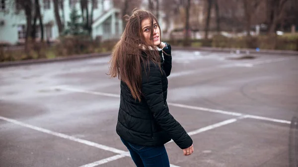 A fiatal gyönyörű modell lány mosolyog és visszanéz a városba. Dinamikusan fiatal lány sétál az utcán.Haj repked a szélben. — Stock Fotó