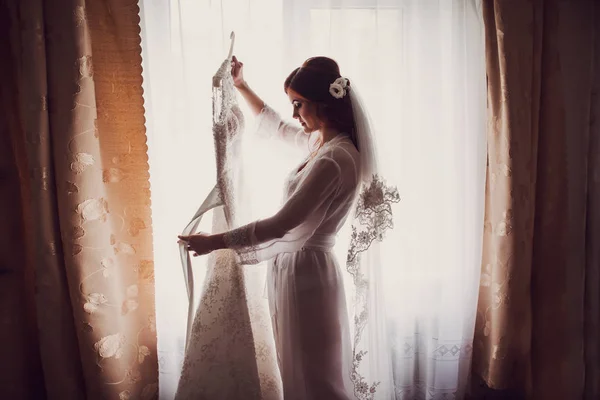 美しさの花嫁が朝に窓の近く彼女のドレスを持っています。 — ストック写真
