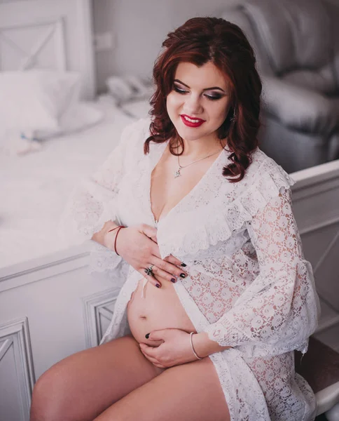 Mujer embarazada feliz sentada en la cama y tocando su vientre en casa —  Fotos de Stock