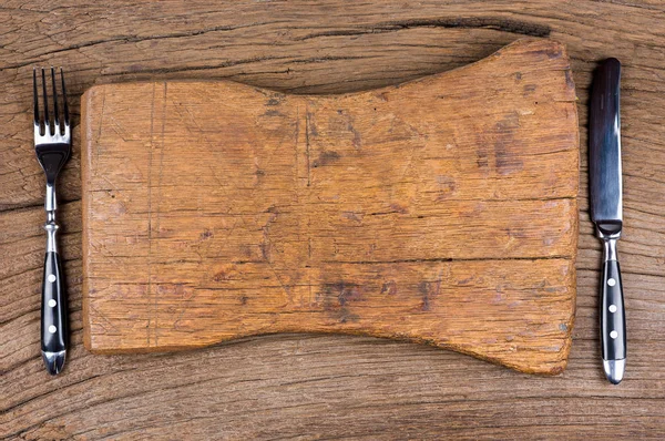 Knife and fork lie together with a very old cutting board on a rustic wood background — Stock Photo, Image