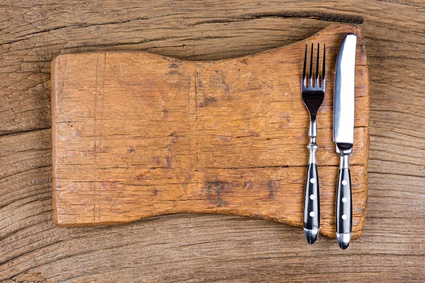 Cuchillo y tenedor se encuentran junto con una tabla de cortar muy antigua sobre un fondo de madera rústica — Foto de Stock