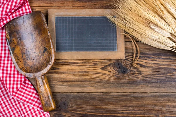 Um quadro-negro de ardósia velho, uma colher de farinha muito antiga e orelhas de grão estão em um fundo de madeira rústica com uma toalha de mesa quadriculada branca vermelha com espaço de cópia — Fotografia de Stock