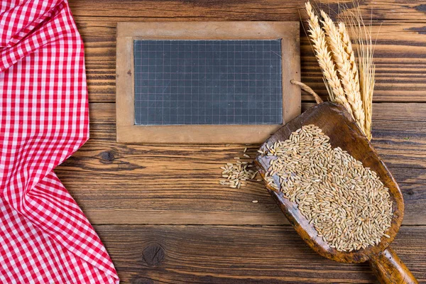 Un vieux tableau noir en ardoise, une très vieille cuillère à farine avec des grains de céréales et des épis de grain reposent sur un fond de bois rustique avec une nappe à carreaux rouges blancs avec espace de copie — Photo