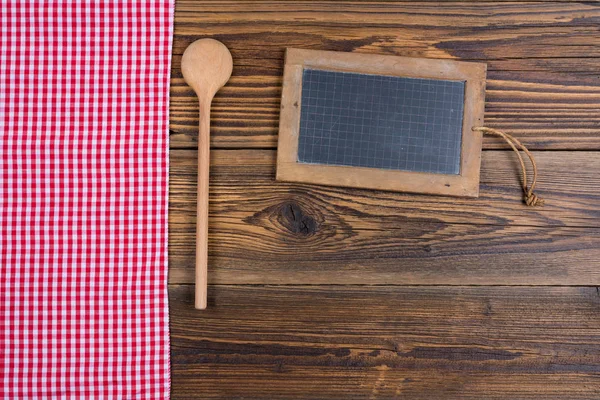 Vecchio sfondo in legno rustico con un cucchiaio da cucina in legno e una vecchia lavagna in ardesia. Sul lato sinistro sono un panno a quadretti bianco rosso. Con spazio di copia sulla lavagna di ardesia e nella parte inferiore dell'immagine — Foto Stock