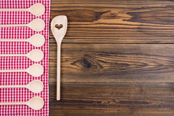 Old rustic wooden background with red white checkered cloth. On the cloth are wooden cooking spoons in a row. With copy space in the right area of the picture — Stock Photo, Image