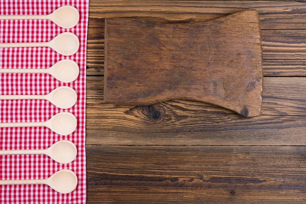 Vecchio sfondo in legno rustico con panno a quadretti bianco rosso. Sul panno ci sono cucchiai da cucina in legno di fila. Con spazio di copia su un vecchio tagliere nella giusta area dell'immagine — Foto Stock