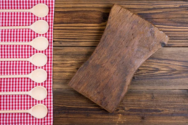 Antiguo fondo rústico de madera con tela a cuadros roja blanca. En el paño hay cucharas de cocción de madera en una fila. Con espacio de copia en una vieja tabla de cortar en el área derecha de la imagen —  Fotos de Stock