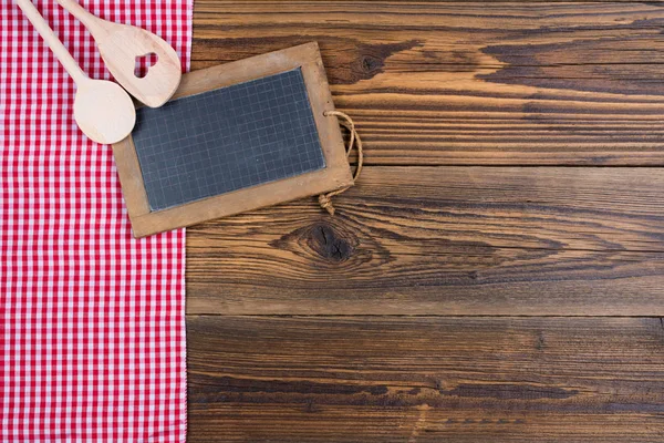 Um velho quadro-negro de ardósia e duas colheres de cozinha de madeira no fundo de madeira rústica no lado esquerdo encontra-se um pano xadrez branco vermelho — Fotografia de Stock