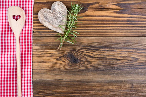 Un cucchiaio di legno con un ritaglio a forma di cuore, rametti di rosmarino e un cuore di legno si trovano su un panno a quadretti rossi e bianchi su uno sfondo di legno rustico con spazio per copiare — Foto Stock