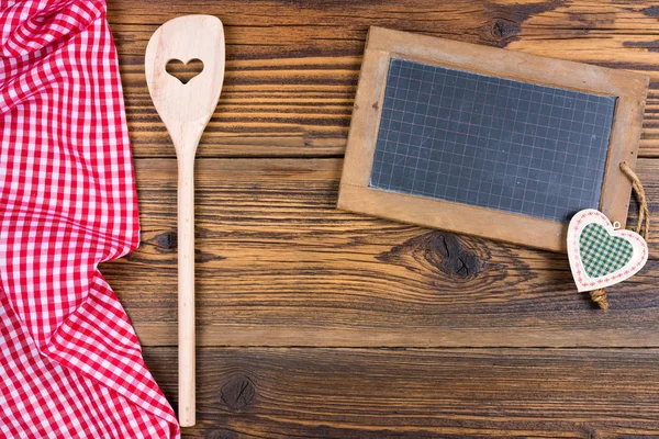 Una vecchia lavagna in ardesia e un cucchiaio da cucina in legno su fondo rustico in legno sul lato sinistro giace un panno a quadretti bianco rosso — Foto Stock