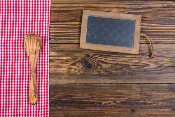 Old rustic wooden background with a wooden cooking spoon and an old slate blackboard. On left side are a red white checkered cloth. With copy space on the slate blackboard and in the lower area of the picture — Stock Photo, Image