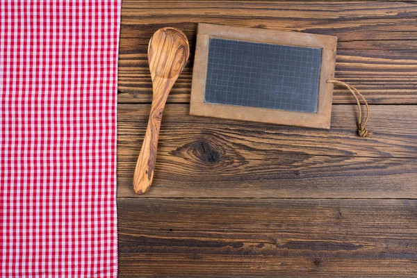 Gammel rustik træ baggrund med en træ madlavning ske og en gammel skifer tavle tavle. På venstre side er en rød hvid ternet klud. Med kopiplads på tavlen tavle og i det nederste område af billedet - Stock-foto