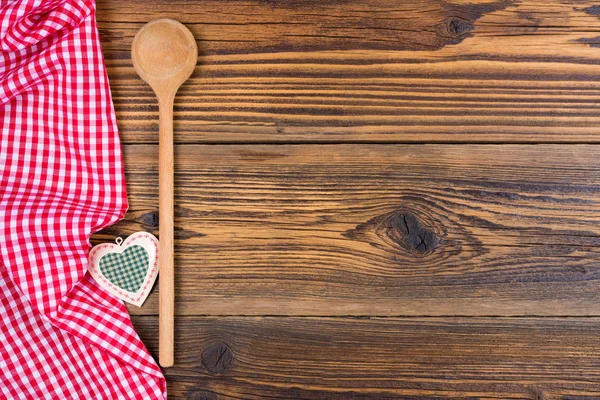 Uma colher de cozinha de madeira velha e coração pequeno da decoração encontra-se em um fundo de madeira rústico com um pano checkered branco vermelho no lado esquerdo — Fotografia de Stock