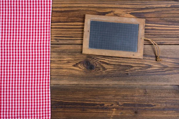 An old slate blackboard lies on a rustic brown wooden background on the left side lies a red white checkered cloth — Stock Photo, Image