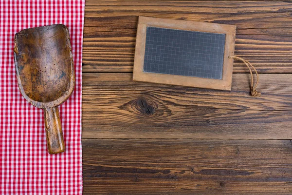 Una vecchia lavagna di ardesia e un vecchio misurino di farina di legno si trova su uno sfondo di legno marrone rustico sul lato sinistro sono un panno a quadretti bianco rosso — Foto Stock