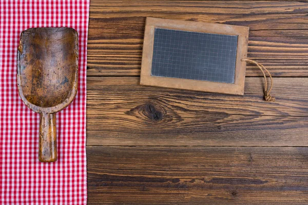 Um velho quadro negro de ardósia e uma velha colher de farinha de madeira repousa sobre um fundo de madeira marrom rústico no lado esquerdo são um pano xadrez branco vermelho — Fotografia de Stock