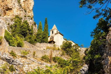 Yaz parlak mavi gökyüzü önünde tarihi dağ köyü Moustiers-Sainte-Marie, Güney Fransa'da kayalıkbir platonun üzerinde dibinde eski şapeli Notre Dame de Beauvoir kadar görüntülemek