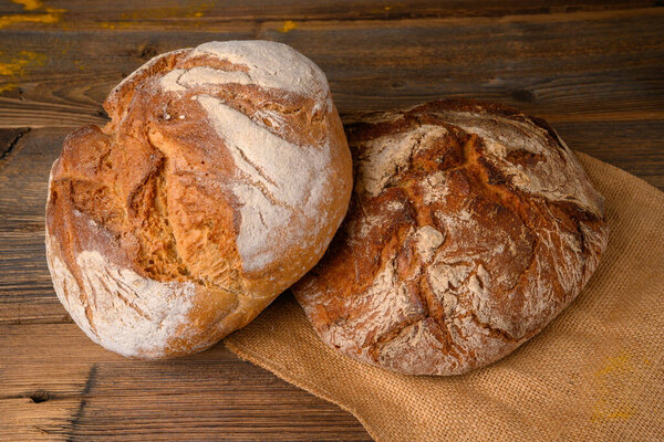 Two fresh whole grain breads from the baker on a jute fabric on a rustic wooden background.