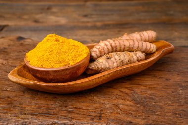 Fresh  whole turmeric roots and dried curcuma powder in a wooden bowl in front of a wooden rustic background.