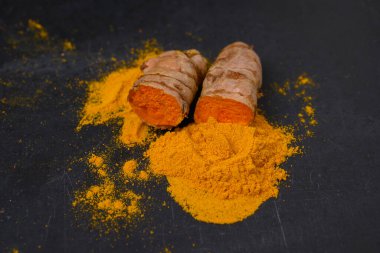 A halved fresh turmeric root and dried curcuma powder on a black slate plate background.