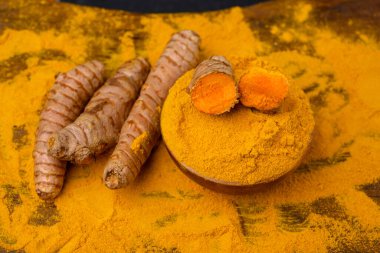 Fresh whole and sliced turmeric roots and dried curcuma powder in a wooden bowl in front of a wooden rustic background.