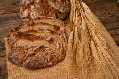 Freshly whole grain bread and bread slices on a jute fabric placed on a rustic wooden background with grain ears.