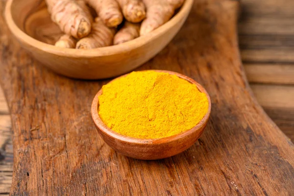 A wooden bowl with curcuma powder and a bowl with whole turmeric roots on a wooden cutting board.