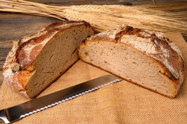 One fresh whole grain bread from the baker, halved with a bread knife on a jute fabric with grain ears on a rustic wooden background.